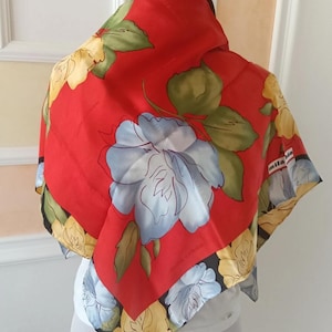 Puede incluir: Un pañuelo de seda rojo con un diseño floral. El pañuelo presenta grandes flores azules y blancas con hojas verdes, y flores amarillas en el borde. El texto "mila schön" es visible.
