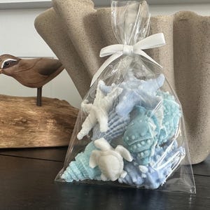 May include: A clear plastic bag tied with a white ribbon containing a collection of small, molded soap figures in shades of light blue and white. The shapes include various seashells, a starfish, and a sea turtle, all with detailed textures.