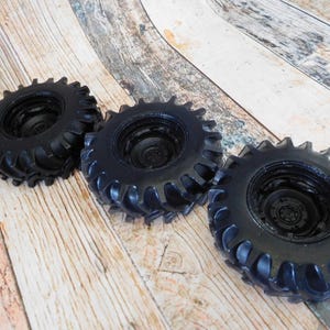 Three handmade monster truck tire shaped soaps in solid black, molded with deep rugged tread patterns and detailed recessed wheel hubs. Two tire soaps lie flat while a third stands upright to show the thick rounded tire shape, chunky off road tread and realistic wheel detailing, displayed against a rustic weathered wood background.