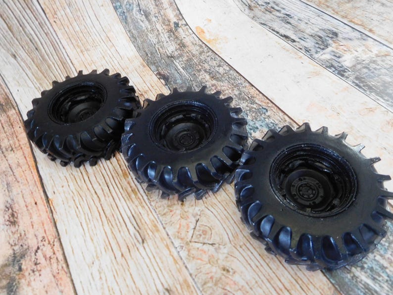 Three handmade monster truck tire shaped soaps in solid black, molded with deep rugged tread patterns and detailed recessed wheel hubs. Two tire soaps lie flat while a third stands upright to show the thick rounded tire shape, chunky off road tread and realistic wheel detailing, displayed against a rustic weathered wood background.