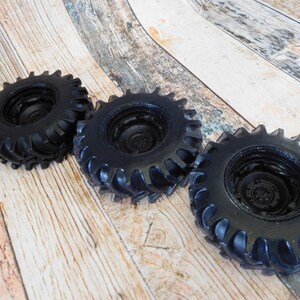 Three handmade monster truck tire shaped soaps in solid black, molded with deep rugged tread patterns and detailed recessed wheel hubs. Two tire soaps lie flat while a third stands upright to show the thick rounded tire shape, chunky off road tread and realistic wheel detailing, displayed against a rustic weathered wood background.