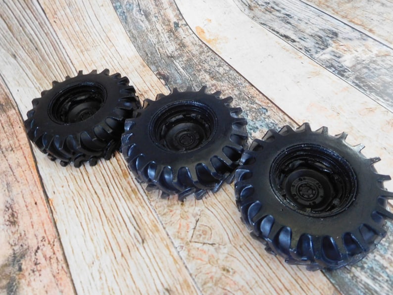 Three handmade monster truck tire shaped soaps in solid black, molded with deep rugged tread patterns and detailed recessed wheel hubs. Two tire soaps lie flat while a third stands upright to show the thick rounded tire shape, chunky off road tread and realistic wheel detailing, displayed against a rustic weathered wood background.