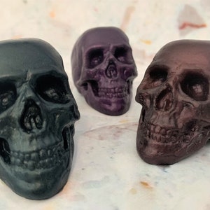 Three translucent skulls sitting in a semi-circle facing each other.  The front left skull is black-green, middle back skull is eggplant purple and front right skull is black-red.