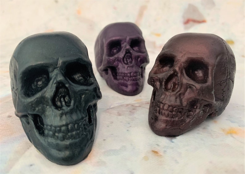 Three translucent skulls sitting in a semi-circle facing each other.  The front left skull is black-green, middle back skull is eggplant purple and front right skull is black-red.