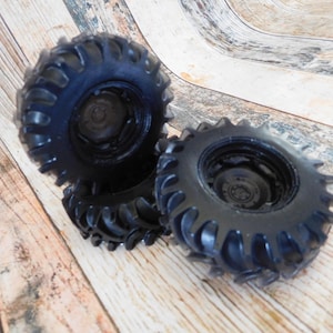 Three handmade monster truck tire shaped soaps in solid black, molded with deep rugged tread patterns and detailed recessed wheel hubs. Two tire soaps lie flat while a third stands upright to show the thick rounded tire shape, chunky off road tread and realistic wheel detailing, displayed against a rustic weathered wood background.