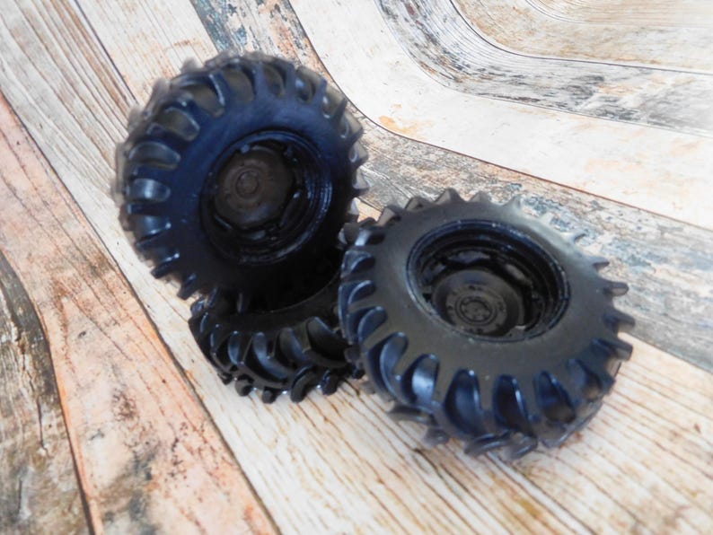 Three handmade monster truck tire shaped soaps in solid black, molded with deep rugged tread patterns and detailed recessed wheel hubs. Two tire soaps lie flat while a third stands upright to show the thick rounded tire shape, chunky off road tread and realistic wheel detailing, displayed against a rustic weathered wood background.