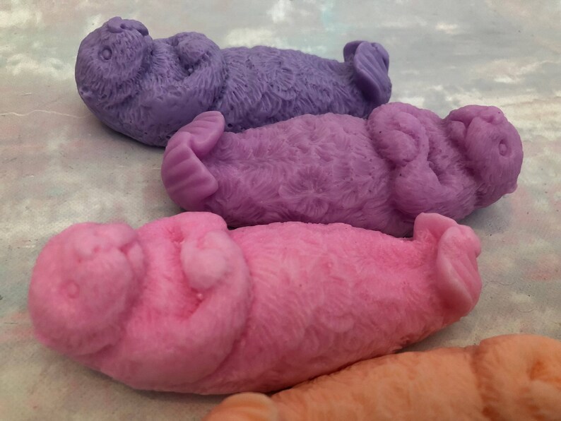 Assorted 3D otter-shaped soaps in black, brown, and soft pastel colors, floating on their backs with arms crossed on their chests and heads and feet facing the sky.