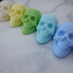 Seven three-dimensional skull shaped soaps in a line, each closer to the lens than the previous. In order from left to right: Black, ivory, yellow, green, blue green, blue, purple.