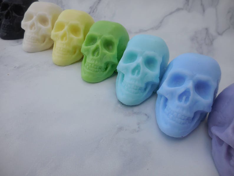 Seven three-dimensional skull shaped soaps in a line, each closer to the lens than the previous. In order from left to right: Black, ivory, yellow, green, blue green, blue, purple.