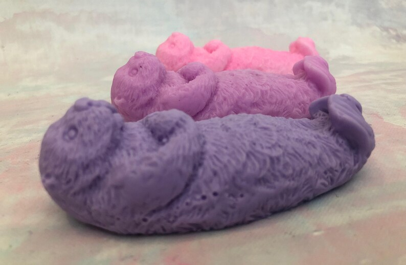 Assorted 3D otter-shaped soaps in black, brown, and soft pastel colors, floating on their backs with arms crossed on their chests and heads and feet facing the sky.