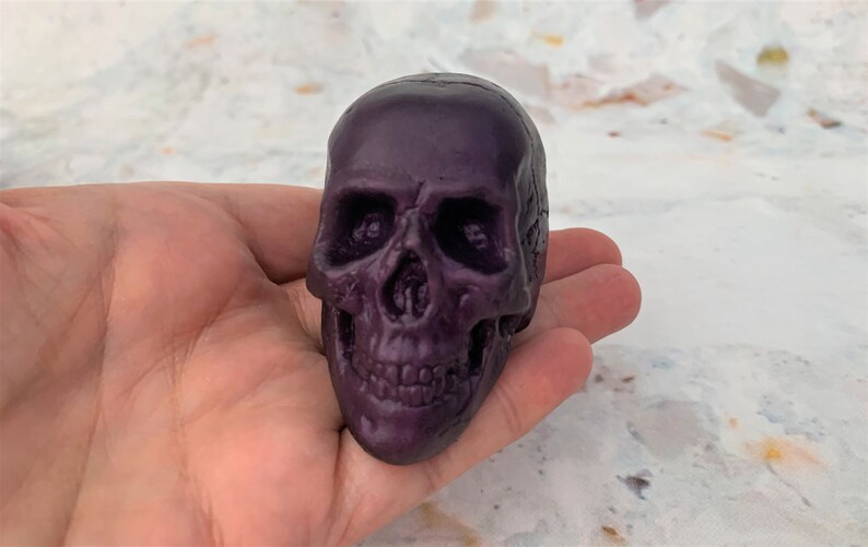 Seller is holding one purple eggplant colored skull atop her fingers with palm facing upwards.  The skull is facing forward.