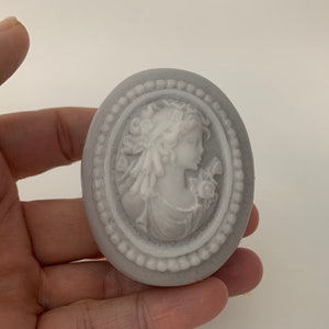 An oval mini soap featuring the illustration of a sophisticated woman facing in one direction, rendered in soft pastel colors of your choice with white highlights that enhance her elegance and charm.  The soap shown is held in someones fingertips
