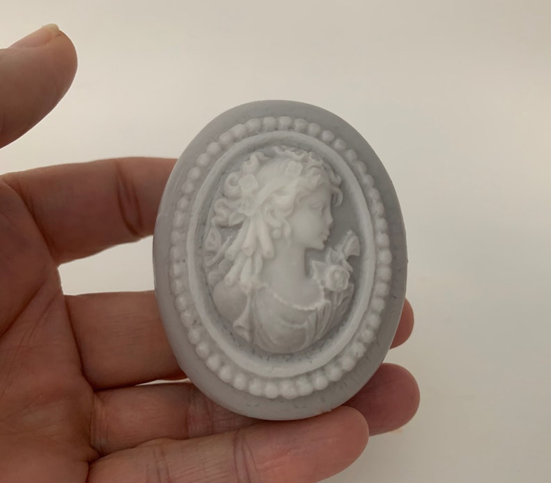 An oval mini soap featuring the illustration of a sophisticated woman facing in one direction, rendered in soft pastel colors of your choice with white highlights that enhance her elegance and charm.  The soap shown is held in someones fingertips