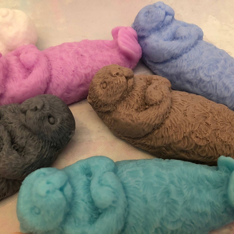 Assorted 3D otter-shaped soaps in black, brown, and soft pastel colors, floating on their backs with arms crossed on their chests and heads and feet facing the sky.