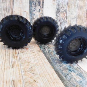 Three handmade monster truck tire shaped soaps in solid black, molded with deep rugged tread patterns and detailed recessed wheel hubs. Two tire soaps lie flat while a third stands upright to show the thick rounded tire shape, chunky off road tread and realistic wheel detailing, displayed against a rustic weathered wood background.