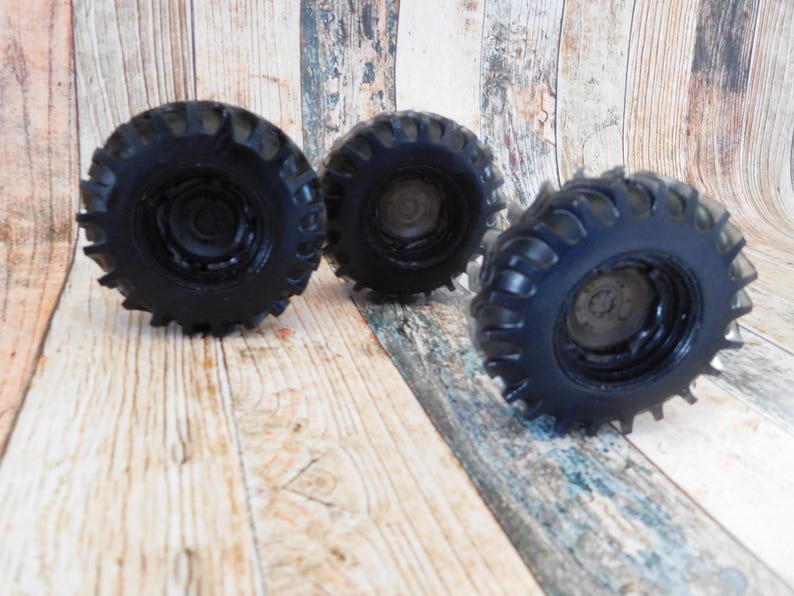 Three handmade monster truck tire shaped soaps in solid black, molded with deep rugged tread patterns and detailed recessed wheel hubs. Two tire soaps lie flat while a third stands upright to show the thick rounded tire shape, chunky off road tread and realistic wheel detailing, displayed against a rustic weathered wood background.