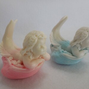 Two beautifully detailed mermaids lie on their bellies, each atop a clam shell. Their arms support their faces while their tails point upwards. A gradient effect adds a touch of enchantment between the white mermaid & the pastel colored clam shell.