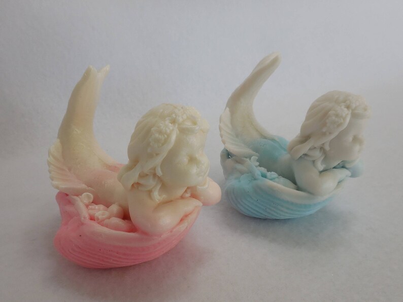 Two beautifully detailed mermaids lie on their bellies, each atop a clam shell. Their arms support their faces while their tails point upwards. A gradient effect adds a touch of enchantment between the white mermaid & the pastel colored clam shell.