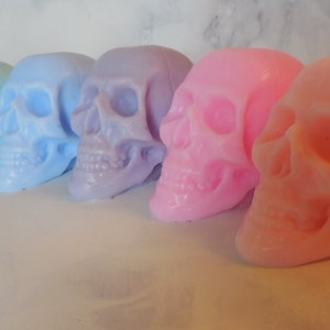 Six three-dimensional skull shaped soaps in a line, each closer to the lens than the previous. In order from left to right: Green, blue green, blue, purple, pink and orange