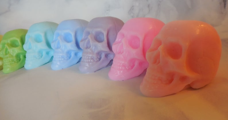 Six three-dimensional skull shaped soaps in a line, each closer to the lens than the previous. In order from left to right: Green, blue green, blue, purple, pink and orange
