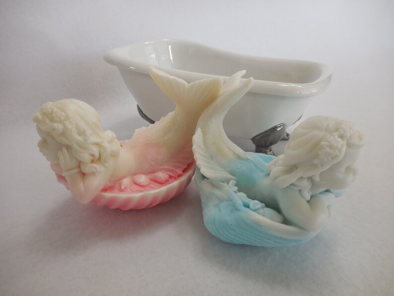 Two beautifully detailed mermaids lie on their bellies, each atop a clam shell. Their arms support their faces while their tails point upwards. A gradient effect adds a touch of enchantment between the white mermaid & the pastel colored clam shell.