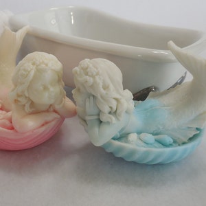 Two beautifully detailed mermaids lie on their bellies, each atop a clam shell. Their arms support their faces while their tails point upwards. A gradient effect adds a touch of enchantment between the white mermaid & the pastel colored clam shell.