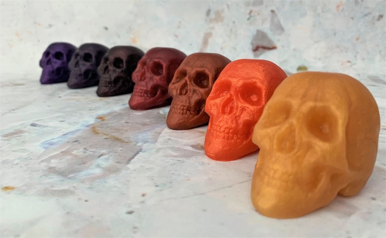 Picture shows 7 skulls in a diagonal row from back left to front right colors are purple galaxy, purple eggplant, black-red, red wine, bronze, orange and gold.