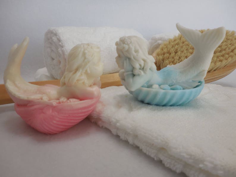 Two beautifully detailed mermaids lie on their bellies, each atop a clam shell. Their arms support their faces while their tails point upwards. A gradient effect adds a touch of enchantment between the white mermaid & the pastel colored clam shell.