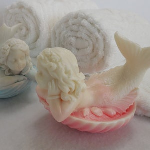 Two beautifully detailed mermaids lie on their bellies, each atop a clam shell. Their arms support their faces while their tails point upwards. A gradient effect adds a touch of enchantment between the white mermaid & the pastel colored clam shell.