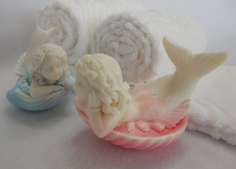 Two beautifully detailed mermaids lie on their bellies, each atop a clam shell. Their arms support their faces while their tails point upwards. A gradient effect adds a touch of enchantment between the white mermaid & the pastel colored clam shell.