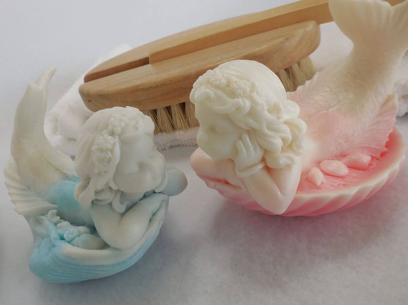 Two beautifully detailed mermaids lie on their bellies, each atop a clam shell. Their arms support their faces while their tails point upwards. A gradient effect adds a touch of enchantment between the white mermaid & the pastel colored clam shell.