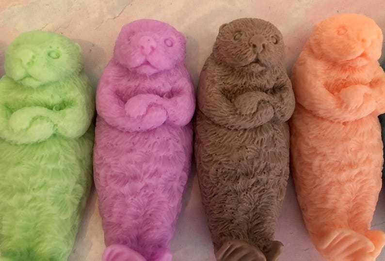 Assorted 3D otter-shaped soaps in black, brown, and soft pastel colors, floating on their backs with arms crossed on their chests and heads and feet facing the sky.