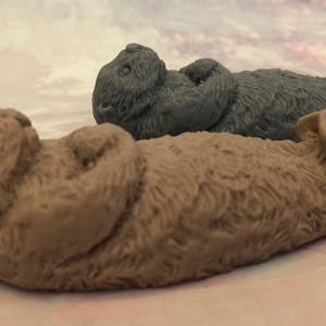 Assorted 3D otter-shaped soaps in black, brown, and soft pastel colors, floating on their backs with arms crossed on their chests and heads and feet facing the sky.