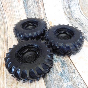 Three handmade monster truck tire shaped soaps in solid black, molded with deep rugged tread patterns and detailed recessed wheel hubs. Two tire soaps lie flat while a third stands upright to show the thick rounded tire shape, chunky off road tread and realistic wheel detailing, displayed against a rustic weathered wood background.
