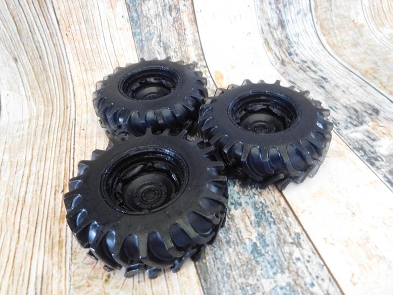 Three handmade monster truck tire shaped soaps in solid black, molded with deep rugged tread patterns and detailed recessed wheel hubs. Two tire soaps lie flat while a third stands upright to show the thick rounded tire shape, chunky off road tread and realistic wheel detailing, displayed against a rustic weathered wood background.
