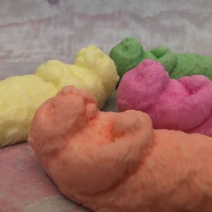 Assorted 3D otter-shaped soaps in black, brown, and soft pastel colors, floating on their backs with arms crossed on their chests and heads and feet facing the sky.