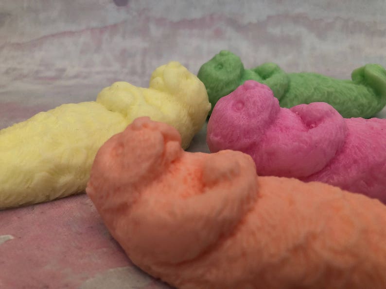 Assorted 3D otter-shaped soaps in black, brown, and soft pastel colors, floating on their backs with arms crossed on their chests and heads and feet facing the sky.