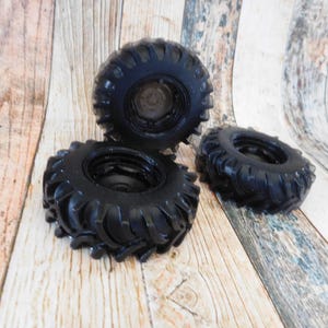 Three handmade monster truck tire shaped soaps in solid black, molded with deep rugged tread patterns and detailed recessed wheel hubs. Two tire soaps lie flat while a third stands upright to show the thick rounded tire shape, chunky off road tread and realistic wheel detailing, displayed against a rustic weathered wood background.
