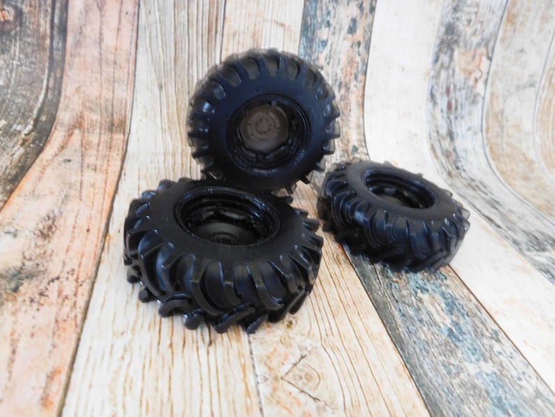 Three handmade monster truck tire shaped soaps in solid black, molded with deep rugged tread patterns and detailed recessed wheel hubs. Two tire soaps lie flat while a third stands upright to show the thick rounded tire shape, chunky off road tread and realistic wheel detailing, displayed against a rustic weathered wood background.