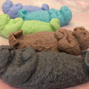 Assorted 3D otter-shaped soaps in black, brown, and soft pastel colors, floating on their backs with arms crossed on their chests and heads and feet facing the sky.