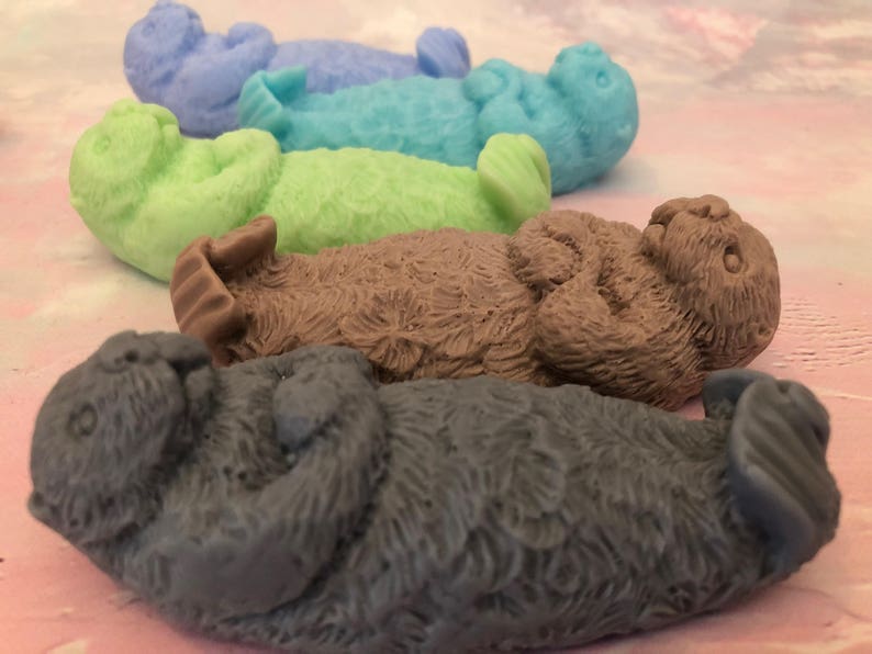 Assorted 3D otter-shaped soaps in black, brown, and soft pastel colors, floating on their backs with arms crossed on their chests and heads and feet facing the sky.