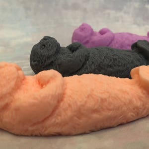 Assorted 3D otter-shaped soaps in black, brown, and soft pastel colors, floating on their backs with arms crossed on their chests and heads and feet facing the sky.