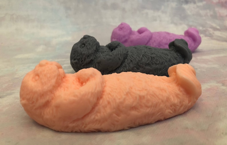 Assorted 3D otter-shaped soaps in black, brown, and soft pastel colors, floating on their backs with arms crossed on their chests and heads and feet facing the sky.