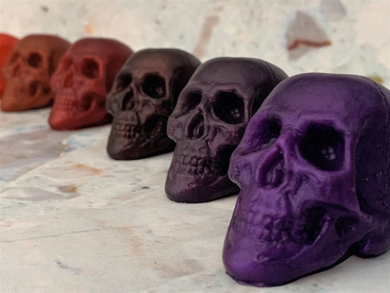 From top left to front right, a horizontal row of translucent skulls in different gradient colors.  Colors from the front right to back left are purple galaxy, eggplant purple, black-red, red wine, bronze, and orange.