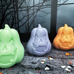 May include: Three colorful Halloween pumpkin decorations. The pumpkins are green, lavender, and orange, each with a carved jack-o'-lantern face. They are placed on a wooden surface with scattered Halloween sprinkles.