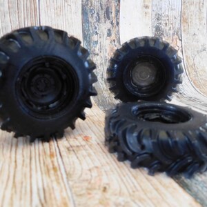 Three handmade monster truck tire shaped soaps in solid black, molded with deep rugged tread patterns and detailed recessed wheel hubs. Two tire soaps lie flat while a third stands upright to show the thick rounded tire shape, chunky off road tread and realistic wheel detailing, displayed against a rustic weathered wood background.