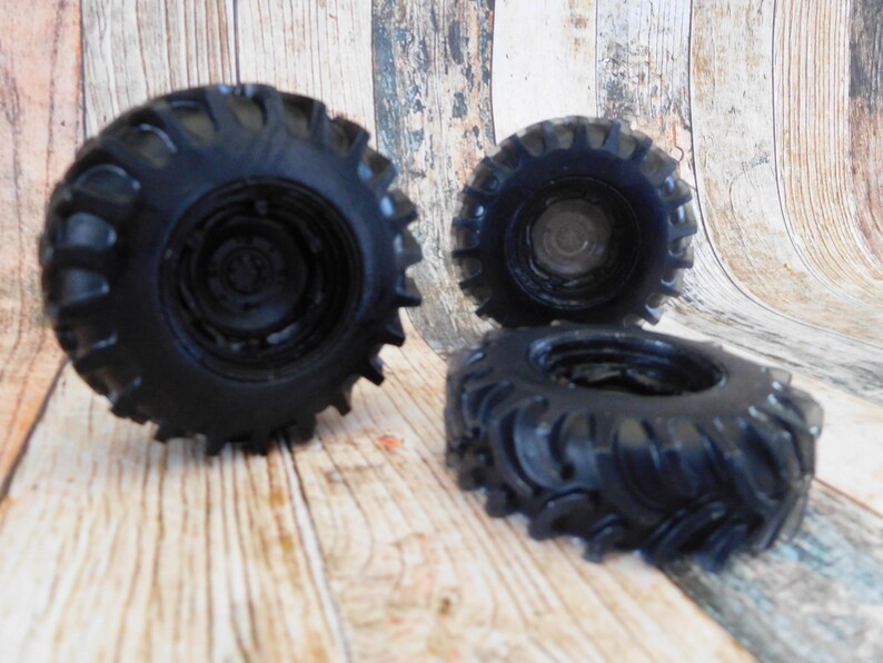 Three handmade monster truck tire shaped soaps in solid black, molded with deep rugged tread patterns and detailed recessed wheel hubs. Two tire soaps lie flat while a third stands upright to show the thick rounded tire shape, chunky off road tread and realistic wheel detailing, displayed against a rustic weathered wood background.