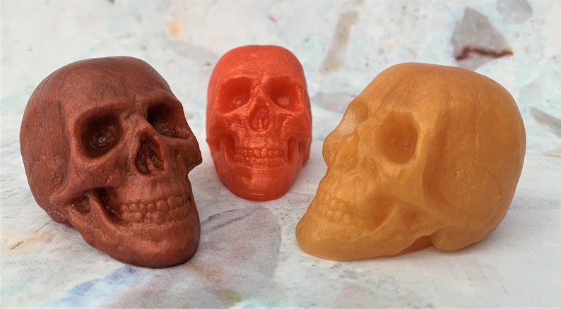 Three translucent skulls sitting in a semi circle facing each other.  Left skull is bronze, back middle skull is orange and right skull is gold.