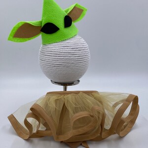 May include: A green felt hat with large ears and black eye accents sits atop a white sphere. Below, a tan tulle skirt with a brown trim and bow detail is displayed. The costume is designed for a pet.