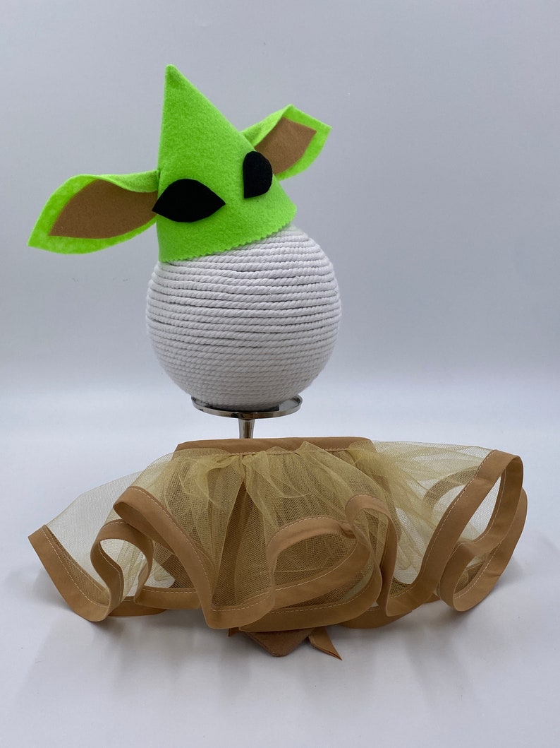 May include: A green felt hat with large ears and black eye accents sits atop a white sphere. Below, a tan tulle skirt with a brown trim and bow detail is displayed. The costume is designed for a pet.